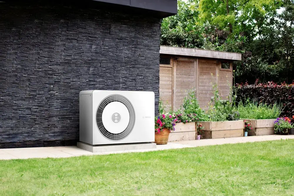 An outdoor heat pump unit, known for its energy efficiency, is installed beside a modern black brick wall, adjacent to a garden with raised wooden planter boxes filled with plants and flowers, on a neatly manicured lawn.